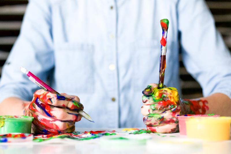 a painter's paint covered hands at work on a canvas, holding brushes in both hands.
